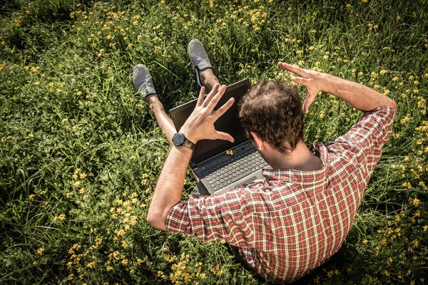 Young man sitting on green grass, upset as something is wrong with his laptop