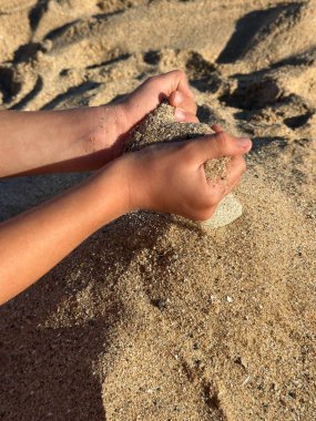 A close-up of small hands cupping warm beach sand with the shoreline in the background. The image conveys a carefree summer mood, childhood curiosity, and the simple joys of seaside vacations.