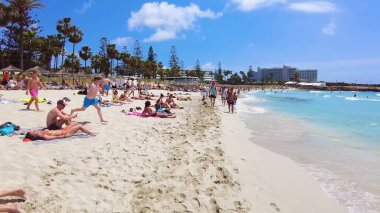 İnsanlar tatil tatili, tur, seyahat, yüzme, oyun, yürüyüş, yemek ve güneşli gökyüzü ve mavi-beyaz bulutlar altında dinlenme ile yaşam tarzları ve plaj titreşimleri. Ayia Napa Adası, Kıbrıs - 8 Temmuz 2023.