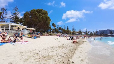 İnsanlar tatil tatili, tur, seyahat, yüzme, oyun, yürüyüş, yemek ve güneşli gökyüzü ve mavi-beyaz bulutlar altında dinlenme ile yaşam tarzları ve plaj titreşimleri. Ayia Napa Adası, Kıbrıs - 8 Temmuz 2023.