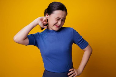 Asyalı kadın kaşınıyor ve parmağını kulaklarına sokuyor sarı arka planda tek başına duruyor..
