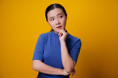 Kafası karışmış Asyalı kadın sarı arka planda tek başına düşünüyor ve ayakta duruyor..