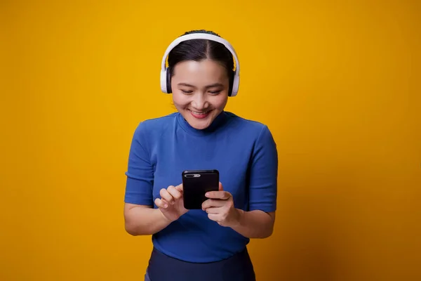 黄色の背景に隔離されたスマートフォンから音楽を聴くヘッドフォンを持つアジアの女性 — ストック写真