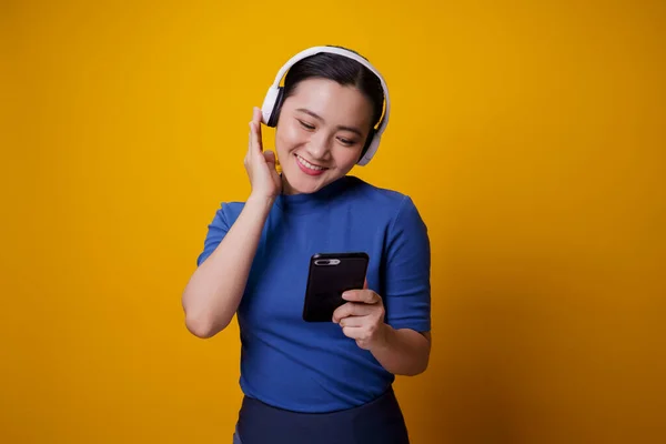 黄色の背景に隔離されたスマートフォンから音楽を聴くヘッドフォンを持つアジアの女性 — ストック写真