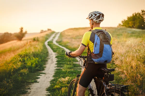 Jovem pedalando em uma estrada rural através do pôr do sol verão prado — Fotografia de Stock