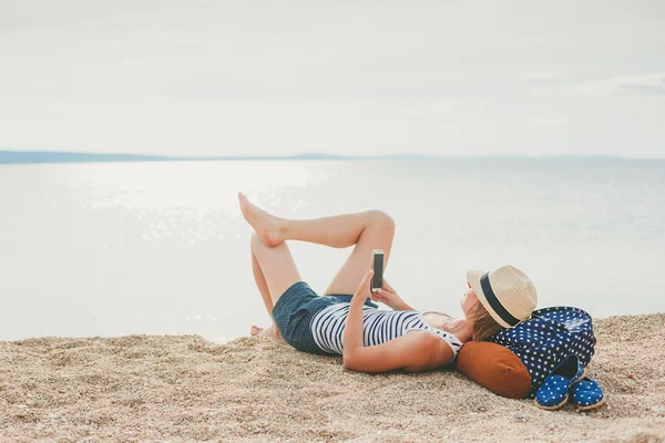 Молода хіпстерська дівчина смс в смартфоні на пляжі з морем на задньому плані — стокове фото