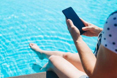 Kadın havuz başında otururken telefonunu kullanıyor.