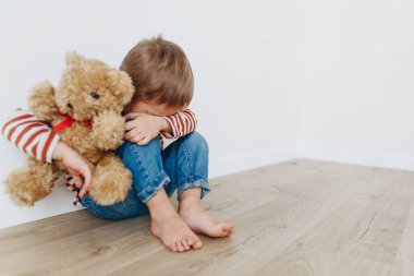 Yalnız ve korkmuş küçük çocuk oyuncak ayıyla boş odada yerde oturuyor.