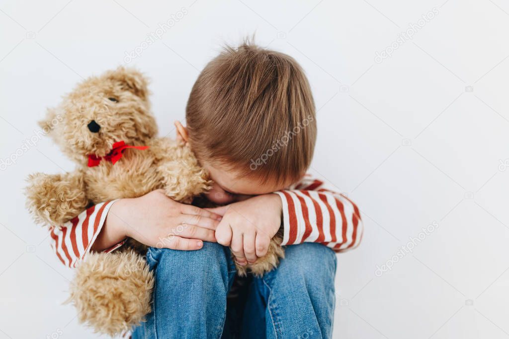 Sad little boy with teddy bear sitting on floor in empty room