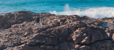 Okyanus dalgaları arkasında parlak mavi gökyüzünün altında engebeli kıyı kayaları üzerinde duran bir adam doğanın gücünü ve deniz kenarında yalnız bir yolcunun huzurunu yakalıyor. Lehior Ojeda 'nın çizgisi.