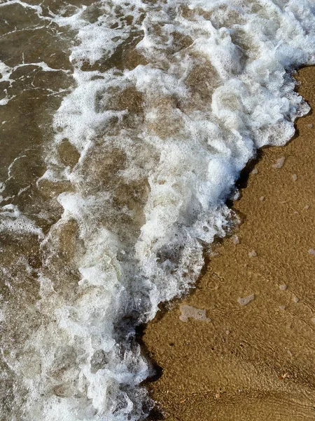 Primer plano de la costa, agua de mar clara con olas poco profundas y una estrecha franja de espuma blanca con burbujas lava la orilla con una superficie lisa de arena marrón —  Fotos de Stock