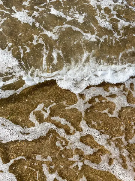 Close-up da costa do mar, água do mar clara com ondas rasas e uma estreita faixa de espuma branca com bolhas lava a costa com uma superfície de areia marrom lisa — Fotografia de Stock