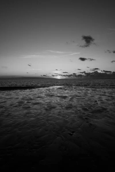 Schwarz-weiß Blick auf die untergehende Sonne, die auf das Meer scheint und sich am Strand spiegelt, Wolken mit sonnenscheinenden Rändern. Landschaft — Stockfoto