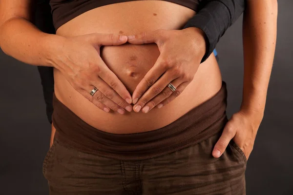 Mani di donna incinta e il suo partner femminile a forma di cuore sul suo ventre — Foto Stock