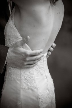 Damat, geline beyaz bir elbiseyle sarılıyor. Sırtı açık, elleri yakın, tanınmaz halde. Siyah beyaz bir fotoğraf.