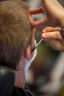 Genç sakallı adam modern berber dükkanında profesyonel olarak saçını kestiriyor. Sakal ve favoriler tıraş olurken ve klasik usturayla şekillendirilirken. Berber dükkanı reklam konsepti. 