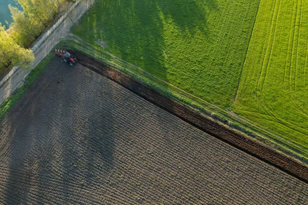 Vista aérea com um drone de um fazendeiro com trator arando uma paisagem rural ondulada de primavera com campos arados e não arados e árvores no céu azul à noite — Fotografia de Stock