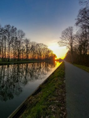 Baharda nehrin üzerinde güzel bir gün batımının manzarası mavi ve altın gökyüzünün arka planında, suyun üzerinde güneş ve ön planda çıplak ağaçların siluetleri.. 