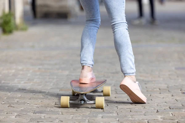 Крупный план женских ног девушки в розовых кроссовках и джинсах на скейтборде на фоне городских улиц — стоковое фото