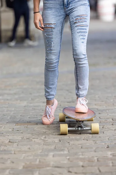 Primer plano de los pies femeninos de una chica con zapatillas de deporte de color rosa y un jeans en el monopatín en el fondo de las calles de la ciudad —  Fotos de Stock