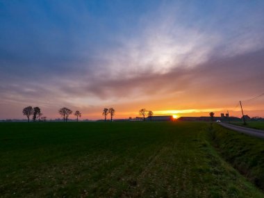 İlkbaharda yeşil çimenler ve sarı çiçekli karahindibalarla kaplı bir arazide dramatik ve renkli bir gün doğumu ya da gün batımı. Güneş ışınları bulutlara doğru ilerliyor..