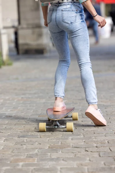 Primer plano de los pies femeninos de una chica con zapatillas de deporte de color rosa y un jeans en el monopatín en el fondo de las calles de la ciudad —  Fotos de Stock