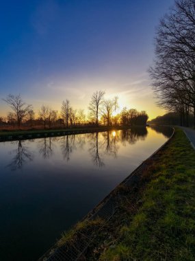 Baharda nehrin üzerinde güzel bir gün batımının manzarası mavi ve altın gökyüzünün arka planında, suyun üzerinde güneş ve ön planda çıplak ağaçların siluetleri.. 