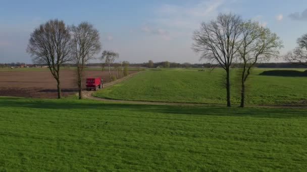 AERIAL strzał drona, Nowoczesny ciągnik ciągnie pustą przyczepę w dół polnej drogi przechodzącej przez pola uprawne. Lecąc w kierunku ciągnika, przemierzając dużą plantację w słonecznej okolicy. — Wideo stockowe