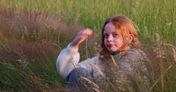 Ładny piękny nastolatek dziewczyna z długimi czerwonymi włosami siedzi na trawie na letnim polu, ciesząc się słońcem i dostosowuje włosy — Wideo stockowe
