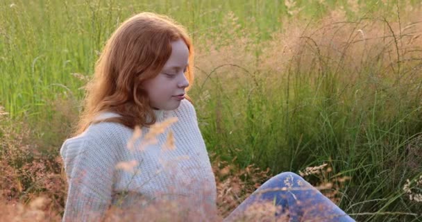 Schattig mooi tiener meisje met lang rood haar zitten op het gras op het zomerveld, genieten van de zon en past haar haar — Stockvideo