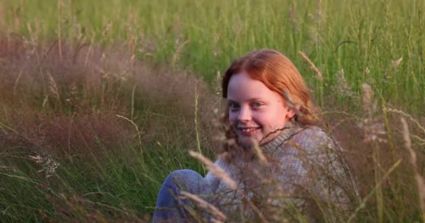Χαριτωμένο όμορφο κορίτσι έφηβος με μακριά κόκκινα μαλλιά κάθεται στο γρασίδι στο καλοκαιρινό πεδίο, απολαμβάνοντας τον ήλιο και προσαρμόζει τα μαλλιά της — Αρχείο Βίντεο