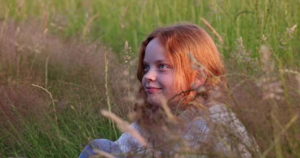 Χαριτωμένο όμορφο κορίτσι έφηβος με μακριά κόκκινα μαλλιά κάθεται στο γρασίδι στο καλοκαιρινό πεδίο, απολαμβάνοντας τον ήλιο και προσαρμόζει τα μαλλιά της σε αργή κίνηση — Αρχείο Βίντεο