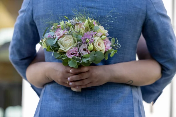 Close-up of a brides hand with a bouquet of white and pink roses that embrace the grooms blue vest on a meadow background — Stock Photo, Image