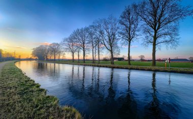 Gün batımında sakin sularda güzel yansıyan yemyeşil ağaçların yer aldığı sakin ve huzurlu bir nehir manzarası.