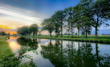 Gerçekten çarpıcı bir gün batımı sakin bir kanalı ışıl ışıl aydınlatıyor, verimli ağaçları ve canlı gökyüzünü mükemmel bir şekilde yansıtıyor.