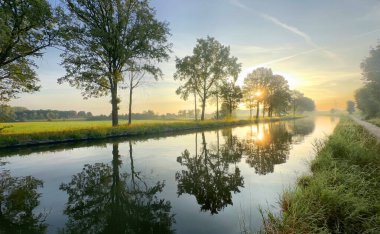 Gün doğarken sakin sulara yansıyan ağaçlarla dolu sakin bir nehir manzarası huzur ve doğa çağrıştırıyor..
