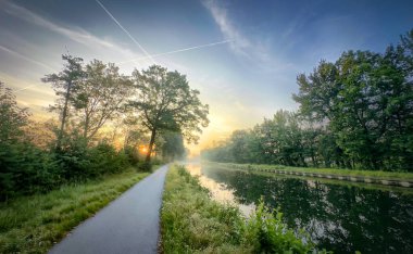 Güneşin doğuşunda dingin ve huzurlu bir nehir yolu, yemyeşil ağaçlar ve yumuşak sabah sisi ile çevrili.