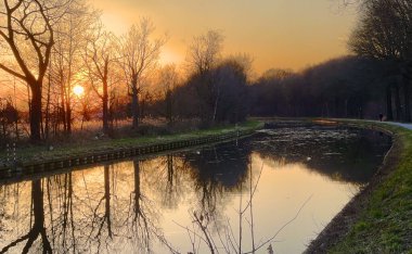 Sakin bir günbatımı, yemyeşil ağaçlar tarafından çerçevelenmiş sakin bir su yolunda canlı yansımaları güzel bir şekilde gözler önüne serer.