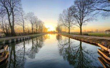 Göz kamaştırıcı ve nefes kesici bir gün doğumu güzel bir şekilde, yemyeşil ağaçlarla çevrili sakin bir kanala yansıyor.