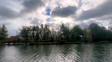 Huzurlu nehir manzarası, bulutlu büyüleyici sahil manzarası, sakin nehir üzerinde ağaçlar ve kulübelerle dramatik bir gökyüzü.