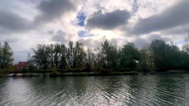 Bulutlu huzurlu deniz kenarı, bulutlu gökyüzünün altında sakin nehir kıyısı, güneş ışığı ve yemyeşil ağaçlarla sakin nehir kenarı manzarası.