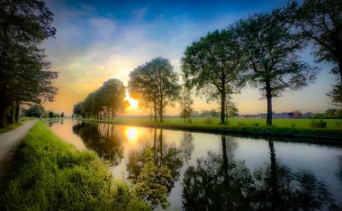 Serin, sakin bir nehir tarafından sersemletici derecede güzel bir gün batımı, yemyeşil ağaçlar ve canlı gökyüzü tarafından mükemmel çerçevelenmiş.