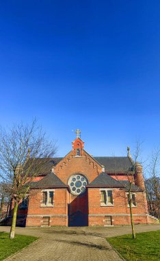 Merksplas, Antwerp, Belçika, 18 Nisan 2025, açık mavi gökyüzünün altında, güzel ağaçlarla çevrili, büyüleyici bir gül penceresi bulunan tuhaf bir kilise.