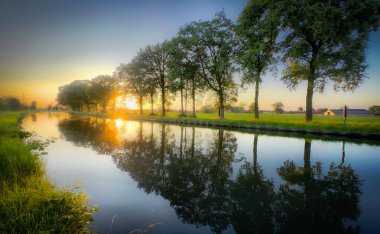 Güzel ve dingin bir günbatımı sahnesi, nehri çevreleyen yemyeşil ağaçlar, durgun suda çarpıcı yansımalar yaratıyor.
