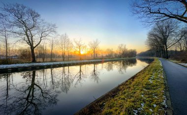 Çarpıcı bir gündoğumu, doğa ve manzara temaları için mükemmel olan sakin bir su yolu boyunca ağaçları aydınlatır.