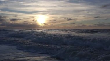 Güneş batan okyanus dalgaları kıyıya doğru yuvarlanıyor, dalgalanan dalgalara yansıyan altın ışık, alçak katmanlı bulutlar gökyüzünde süzülüyor.,
