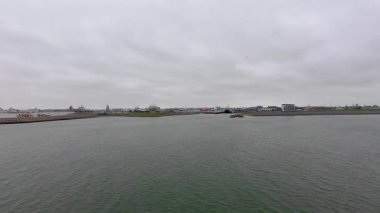 Den Helder, Hollanda, 25 Ekim 2024, sakin su, uzak sanayi limanı, rıhtım gemileri, hafif dalgalar, gri siluet, bulutlu gökyüzü,