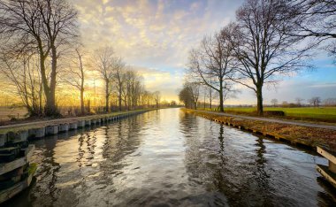 Güzel ve sakin bir su yolu etrafını çevreleyen çıplak ağaçlar tarafından çerçevelenmiş çarpıcı derecede renkli bir günbatımını yansıtır.
