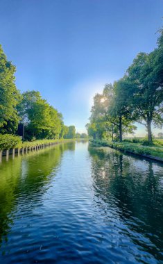 Berrak mavi gökyüzünün altında güzel, sakin bir nehir görülür ve yemyeşil, canlı yeşilliklerle çevrilidir.
