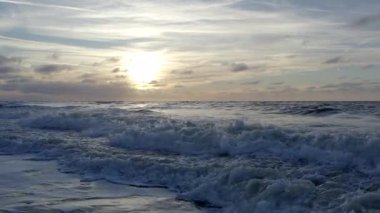 powerful waves crashing into shore, foamy surf exploding under low sun, moody cloud streaks, turbulent water motion, high-energy